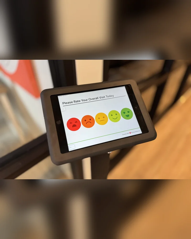 Cafeteria feedback survey kiosk setup: a SurveyStance emoji terminal on a black floor stand beside an exit door.